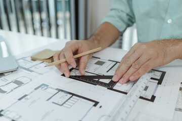An architect uses a metal triangle ruler to draw precise lines on house blueprints laid out on a white desk, focusing on accuracy and detail while working on architectural building plans.