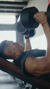 Side view of a young man performing a bench press with dumbbells in a gym, focused and determined. Young Man Doing Weightlifting Bench Press Exercise at the Gym. Vertical