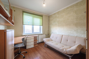 Cozy room featuring a sofa, desk with a rolling chair, wooden flooring, and a window with a roller blind. Wallpaper with a subtle pattern covers the walls