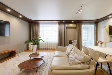 Bright living room with a beige sofa, wooden coffee tables, TV, and plants. Curtains frame a window with natural light. A modern and cozy space