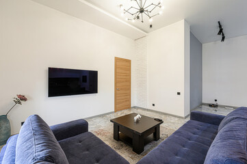 living room with two blue sofas, a dark wood coffee table, a TV, a door, and a modern light fixture. The room is decorated in neutral tones with a patterned rug