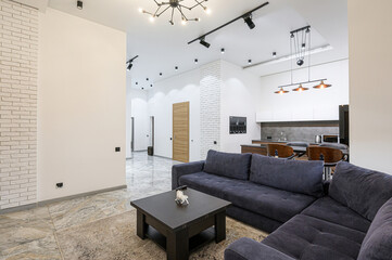 Modern living space with a blue sectional sofa, dark wood coffee table, and open kitchen. White walls with brick accents