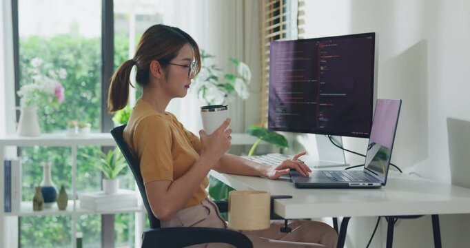 Young Asia female freelance eyeglasses drink coffee and type write prompt AI bot IT app program nomad, video game, terminal with coding, user interface, data center on desktop computer at home office.