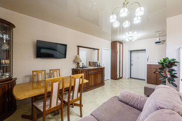 well-lit living/dining area with classic wooden furniture, including a dining table, chairs, and a display cabinet. A large TV is mounted on the wall above the table
