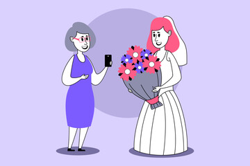 Bride in white gown holding beautiful flower bouquet while smiling bridesmaid taking photo with smartphone