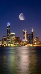 Fototapeta premium Night cityscape reflected in calm water, crescent moon above