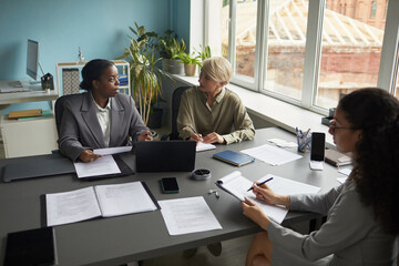 Three diverse businesswomen sitting at table discussing documents and using laptops in modern office meeting room, high angle view