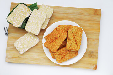 Raw and cooked tempeh displayed side by side on a wooden board, showcasing traditional Indonesian soy-based food prepared and served in a local setting.