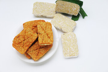 Raw and fried tempeh pieces displayed on a white surface and white plate, highlighting texture and color changes in traditional Indonesian soy-based food.