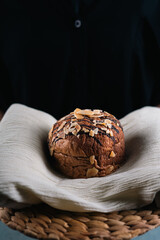 A round chocolate bread topped with almond slices placed on a white cloth and rattan tray, against a dark background.