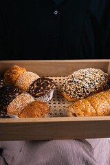A variety of savory rolls with assorted toppings, including sesame, cheese, and chocolate, displayed in a wooden tray on a person’s lap.