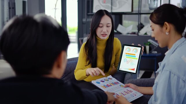 In a modern office, Asian businesswoman presents data summary charts on a tablet to her colleagues, financial report analysis teamwork strategy planning corporate success, marketing, data analytics