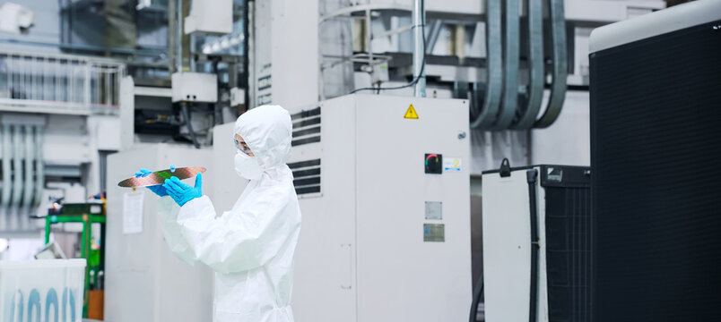 A female worker in a dustproof suit inspecting silicon wafers in a semiconductor factory - Powered by Adobe