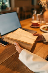 Close-up of woman holding open book in hands at wooden cafй table, blurred laptop and cup in background, cozy studying and learning atmosphere