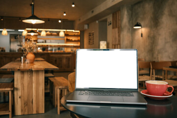 Modern laptop with blank screen and red coffee cup on table in cozy cafe interior, warm light and wooden furniture create workspace atmosphere