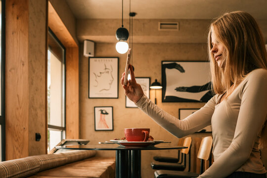 Young woman taking photo with smartphone at cafe table, red coffee cup and breakfast plate in front, modern stylish interior with wall art
