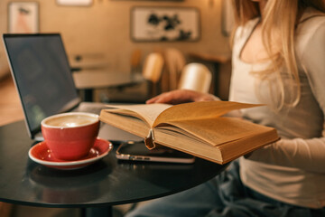 Open book in hands of young woman at cafe table with laptop and red coffee cup, cozy reading and studying atmosphere