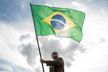 Portrait of cheerful young man looking forward while standing proudly with holding Brasil flag over his head against wind and blue sky, Man holding the Brasil flag celebration