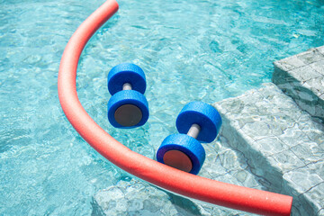 Two pieces of aquatic exercise equipment, a buoyant dumbbell and a swim noodle, are positioned by the edge of a swimming pool. Water-based fitness, hydrotherapy, and rehabilitation exercises.