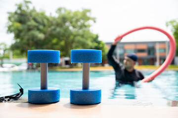A smiling person in a swim cap and rash guard is in a pool, holding a pink swim noodle in an arc above their head. This image depicts aquatic exercise and joyful water recreation.