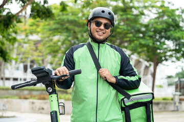 Obraz premium Rider food delivery man wearing green uniform and helmet ride the EV scooter delivery food service to customer. Happy delivery man with green backpack shipping of goods to customers by scooter