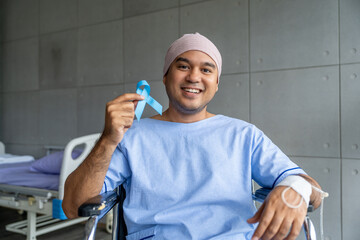 A male cancer patient sits in wheelchair with IV drip holding a blue ribbon symbolizing prostate cancer awareness. Man in treatment holds a blue ribbon representing cancer support hope and advocacy