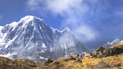 Trekkers trekking on rugged path towards Mt. Annapurna south mountain peak of nepal Himalaya under partial cloud cover. Grassland meadow covered in dry grass in foreground.