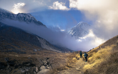 Annapurna base camp trek with trekkers hiking the trail in grassland at sunrise, snow clad Annapurna south peak under cloud cover. Sun rising from behind mountain.