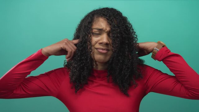 Woman wearing red shirt covers her ears with both hands and grimaces in studio; annoyance frustration.