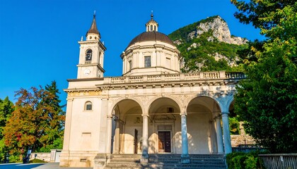 Fototapeta premium A stately, light beige church with a large dome, surrounded by lush green trees and a backdrop of a mountain range, presents a serene and historical architectural scene.