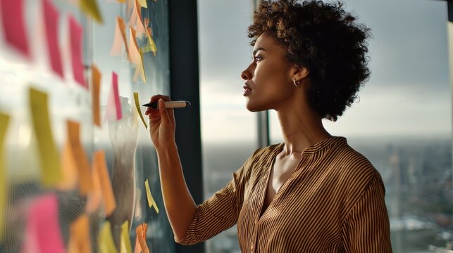 young african american executive manager businesswoman writing strategy ideas on sticky notes on whiteboard doing business project presentation in contemporary corporate office with panoramic view no