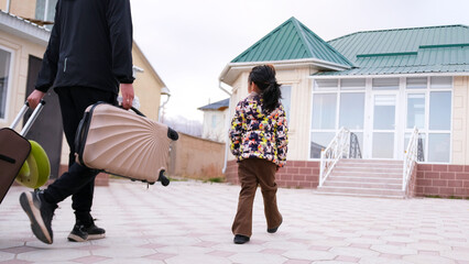 Dad and daughter are returning from vacation. A man with two suitcases and a small child walk along the sidewalk to their house. Traveling with children