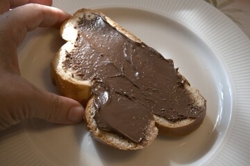 A single slice of sweet bread topped with creamy hazelnut spread served on a white plate with hand in natural morning light. Concept of breakfast, comfort food, and homemade sweetness.