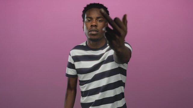Young african american man in a striped shirt flipping middle finger in a pink studio; defiance rebellion.