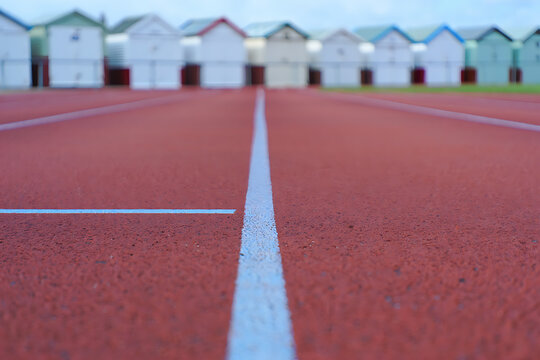 Perspective shot of an empty red running track with white lines and a row of colorful beach huts in the background on a bright day - Powered by Adobe
