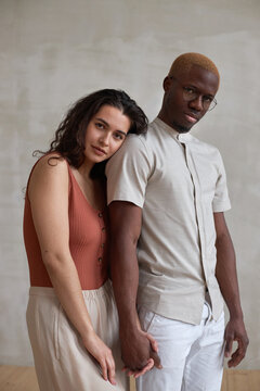 Vertical medium studio portrait of ethnically diverse young couple wearing stylish casual clothes holding hands looking at camera