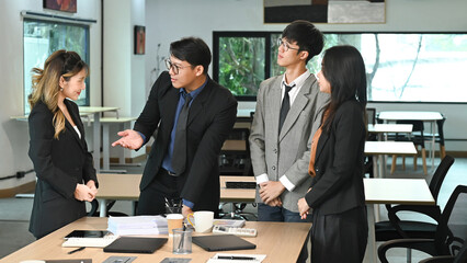 A group of people are stand around a table in a business setting. They are dressed in business attire and appear to be discussing something important