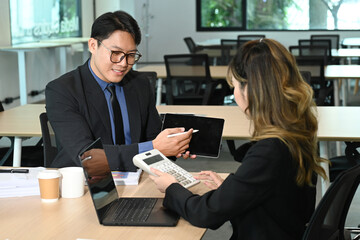 A man and a woman are sitting at a table with a laptop and a tablet. The man is showing the woman a tablet, possibly a tablet for work or personal use.