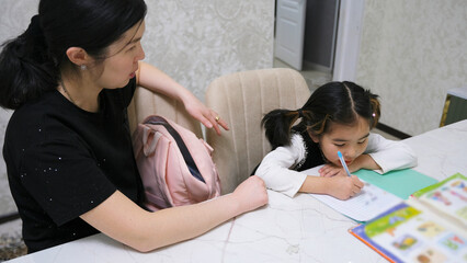 Obraz premium A woman helps a little girl write. The tutor is sitting with the child. Education and training. My daughter is doing her homework