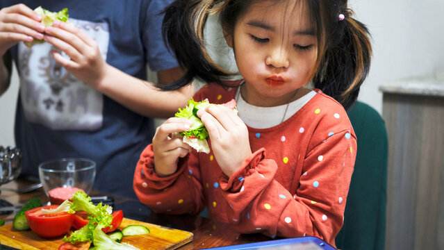 A funny little girl eats a sandwich with slices of sausage, cucumbers and lettuce leaves and watches a cartoon on her tablet