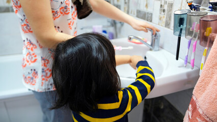 A woman helps a child brush his teeth. A mother and her little daughter are standing at the sink in the bathroom and doing oral hygiene