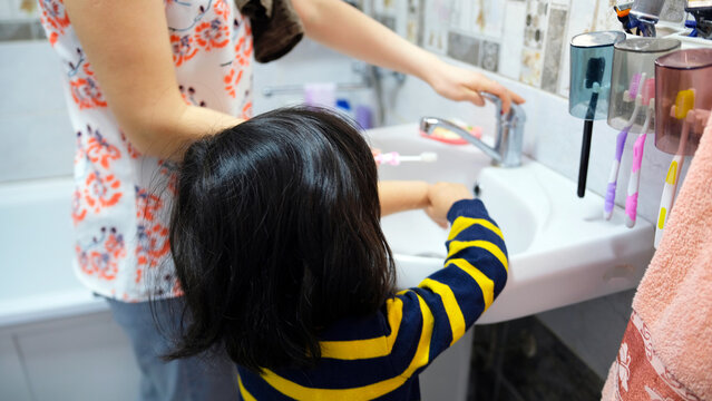 A woman helps a child brush his teeth. A mother and her little daughter are standing at the sink in the bathroom and doing oral hygiene