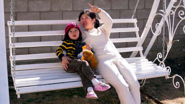 A caring babysitter is rocking with a child on a swing bench. A woman and a child are sitting on a white swing. Mom points at something to her daughter - Powered by Adobe