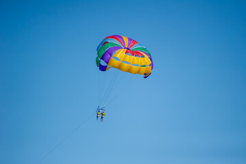 Two people soar high beneath a vibrant parasail against a clear blue sky, capturing the thrill of airborne adventure.