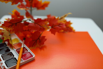 A bright orange canvas is prepared for painting, accompanied by a palette of watercolors and artificial autumn leaves. The scene is set for a creative and colorful activity.