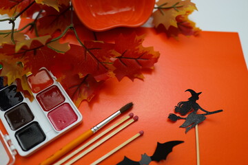 Bright orange backdrop features paint supplies and autumn leaves. Brushes and a witch silhouette on a stick represent Halloween crafting activities in preparation for the festive season.