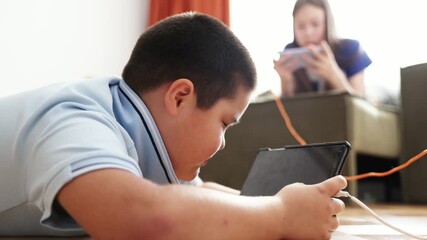 boy lying on wooden floor playing with tablet while girl relaxing on sofa using smartphones with orange curtains, daylight, technology education leisure, horizontal