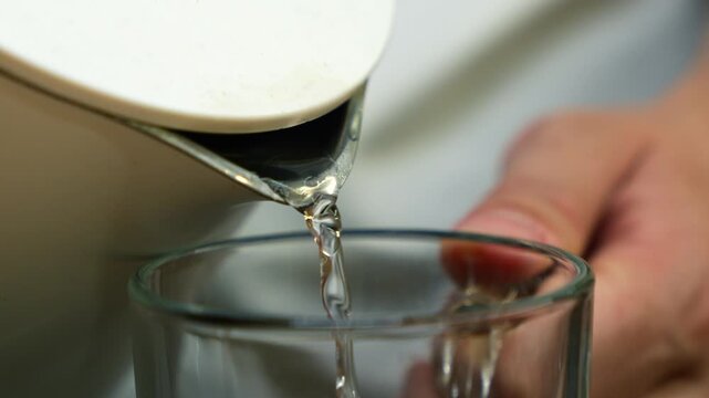 White electric kettle fills empty glass cup with boiled water.
Boiled hot water poured into cup close-up