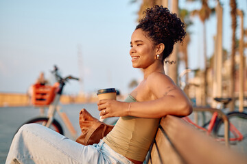 Smiling woman holding disposable coffee cup sitting on bench near bike sharing station, enjoying relaxing moment by the beach