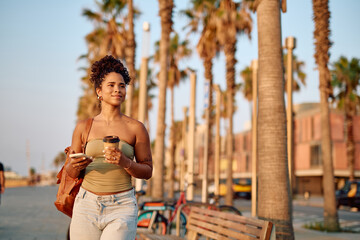 Tourist enjoying a morning walk with takeaway coffee and smartphone along Tel Aviv beach promenade, palm trees and urban background